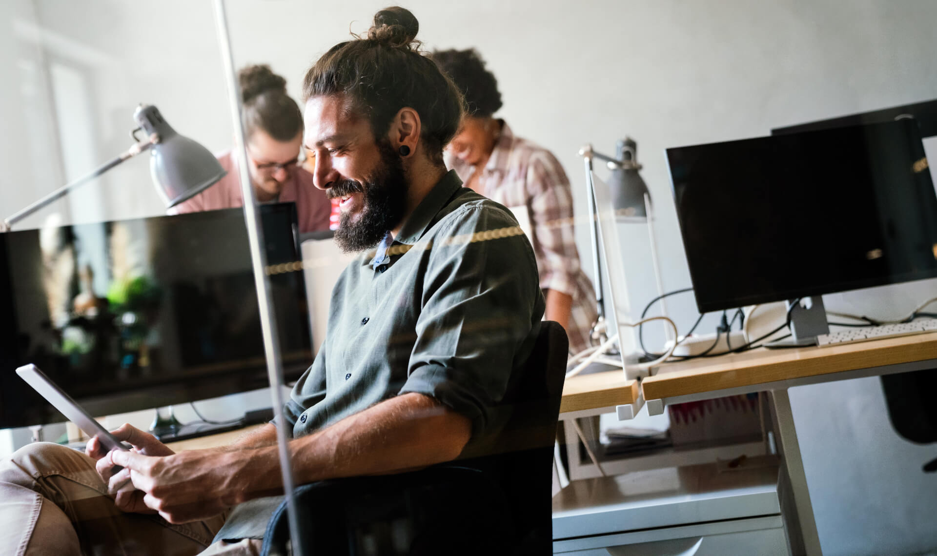 Man smiling at tablet in modern office with two colleagues working at desks behind him, reviewing Veteran Services resources.