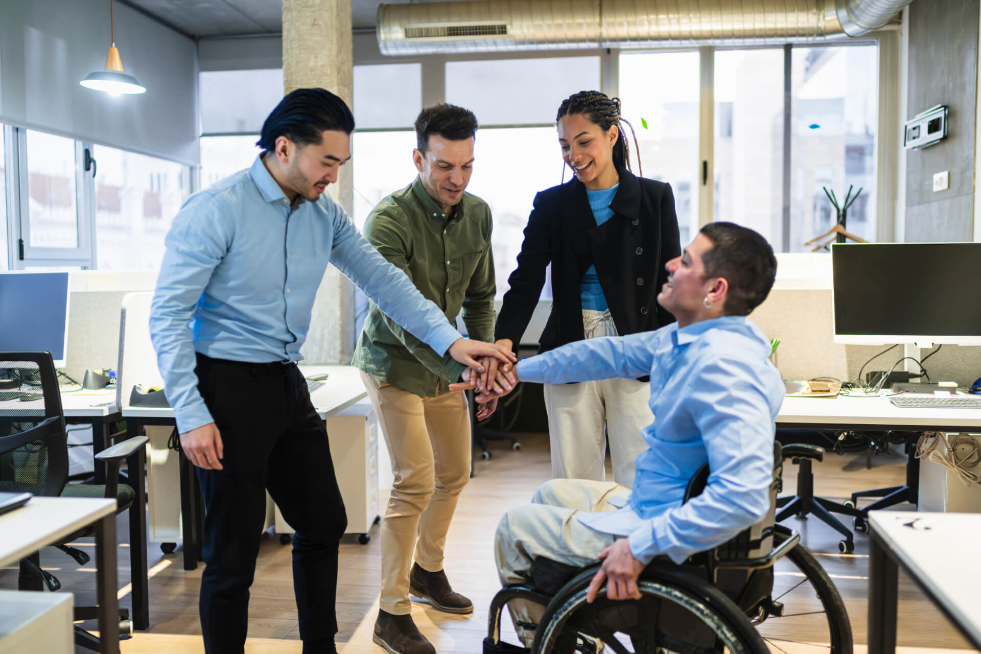 Four diverse coworkers, one in a wheelchair, stack hands in unity at a modern office workspace.