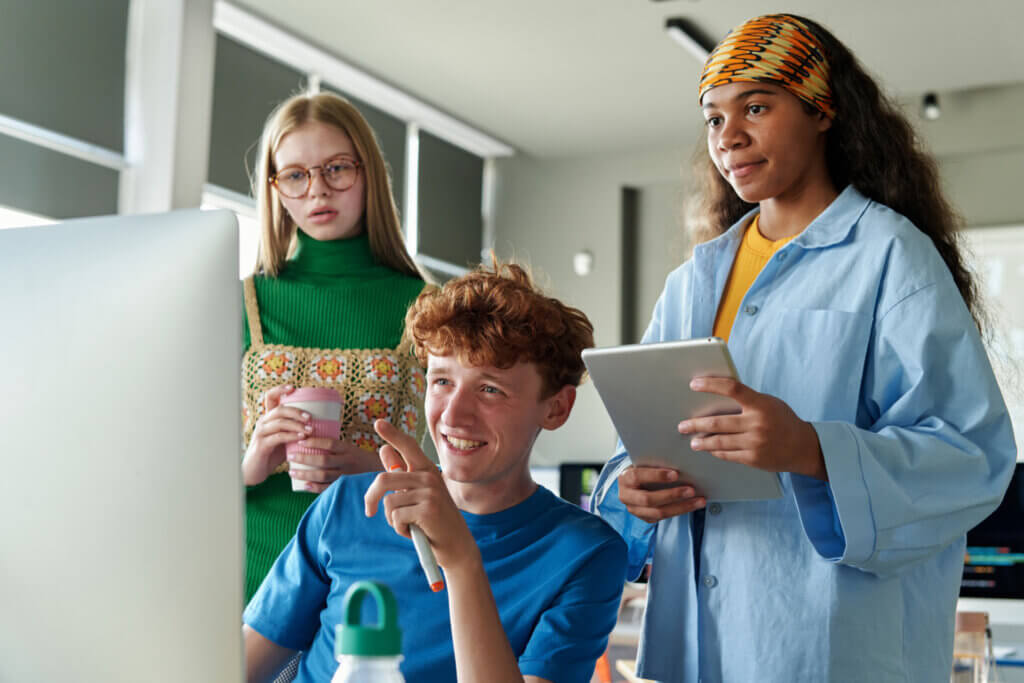 Three young adults look at a computer screen together; one holds a drink, another holds a tablet and smiles.