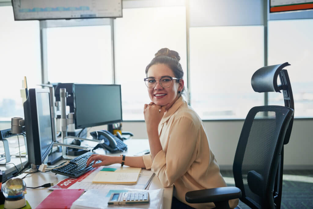 Woman with glasses smiling at her desk in a modern office, surrounded by monitors and office supplies, confidently exploring new career pathways.