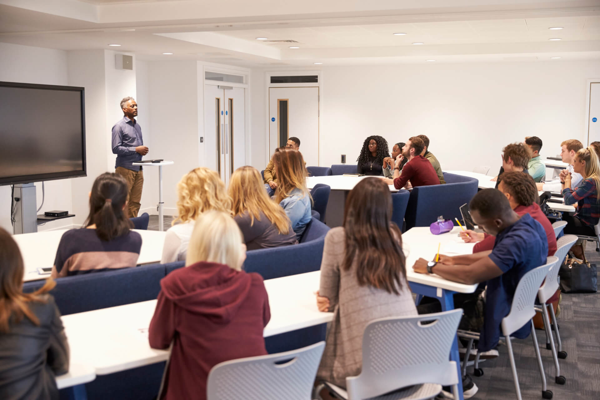 A teacher stands at the front of a classroom speaking to students seated at tables, listening attentively.