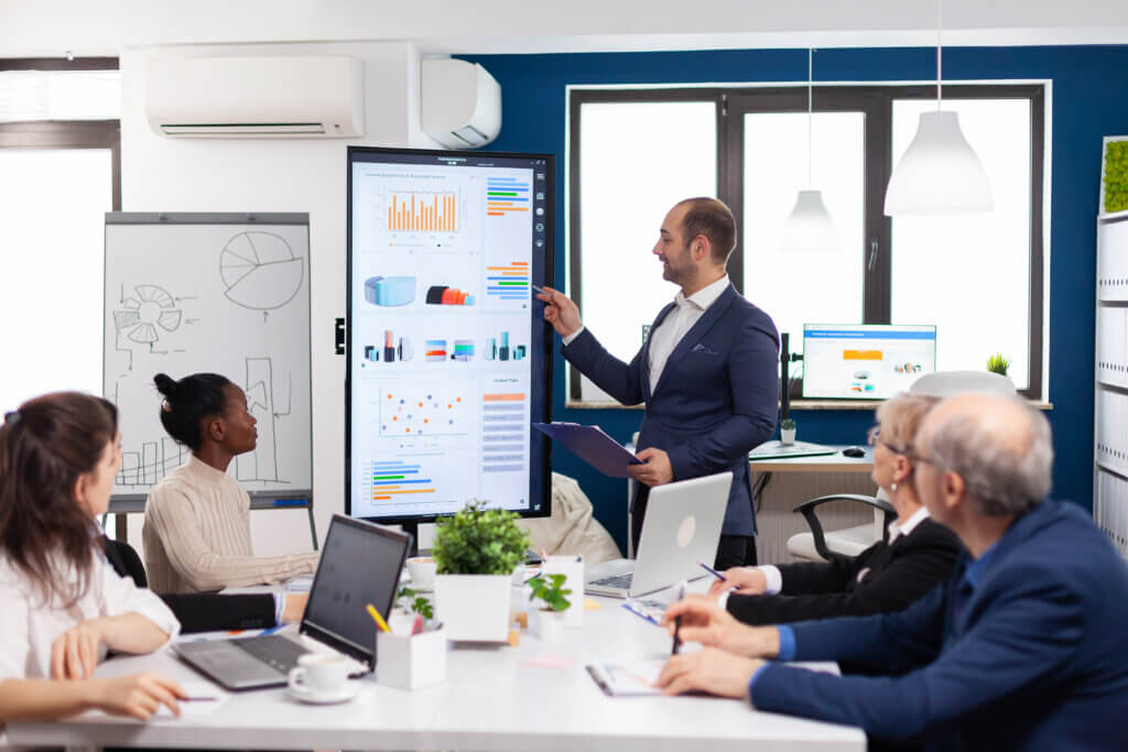 A man presents charts on a large screen during a staff training session to colleagues seated around a conference table with laptops.