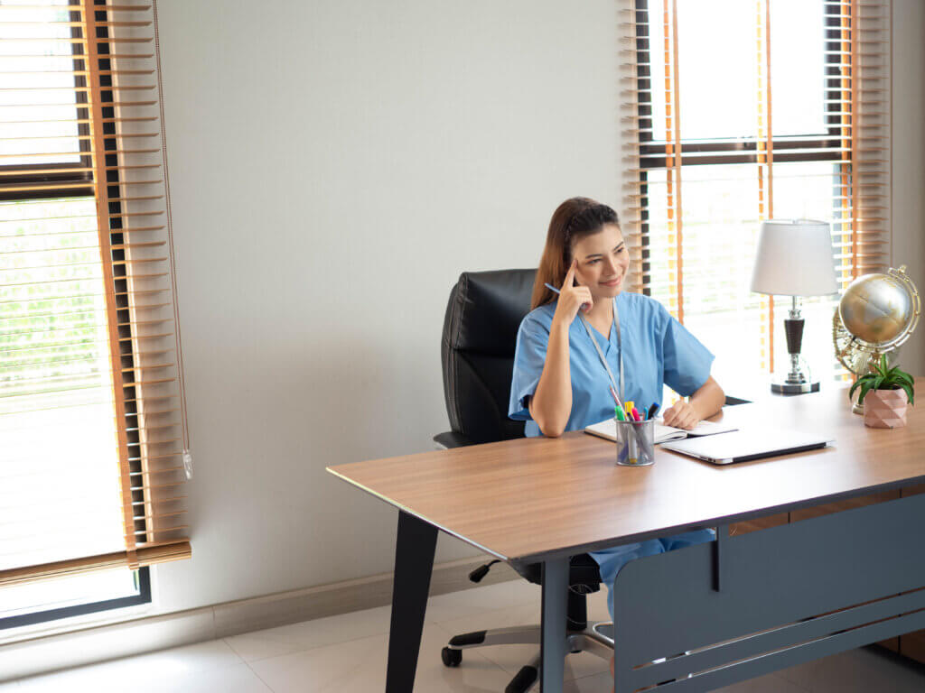 Woman in scrubs sits at an office desk, smiling thoughtfully with her hand on her temple, reflecting on the rewarding careers in healthcare.