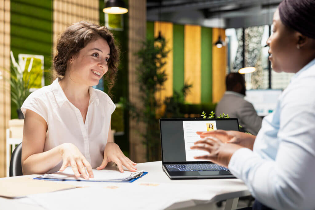 Two women talking at a desk in an office, with a laptop displaying a resume and relevant labor market information on the screen.
