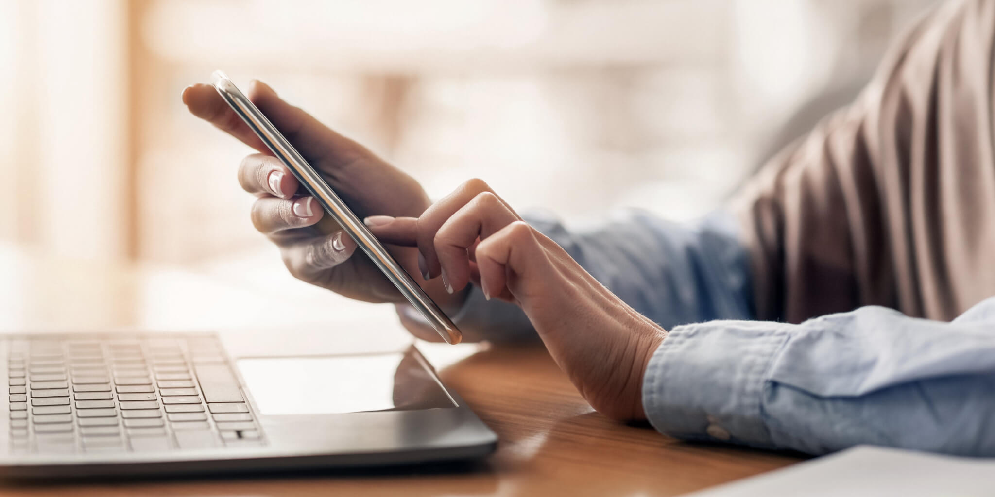 Person using a smartphone while sitting at a desk with an open laptop, wearing a blue shirt, and reviewing labor market insights to stay updated on the latest workforce data.