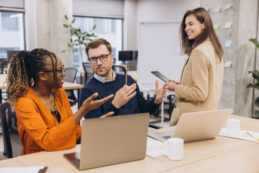 Three colleagues discuss at a desk with laptops in a modern office; one stands holding a tablet, smiling, as they collaborate on Adult Services projects.