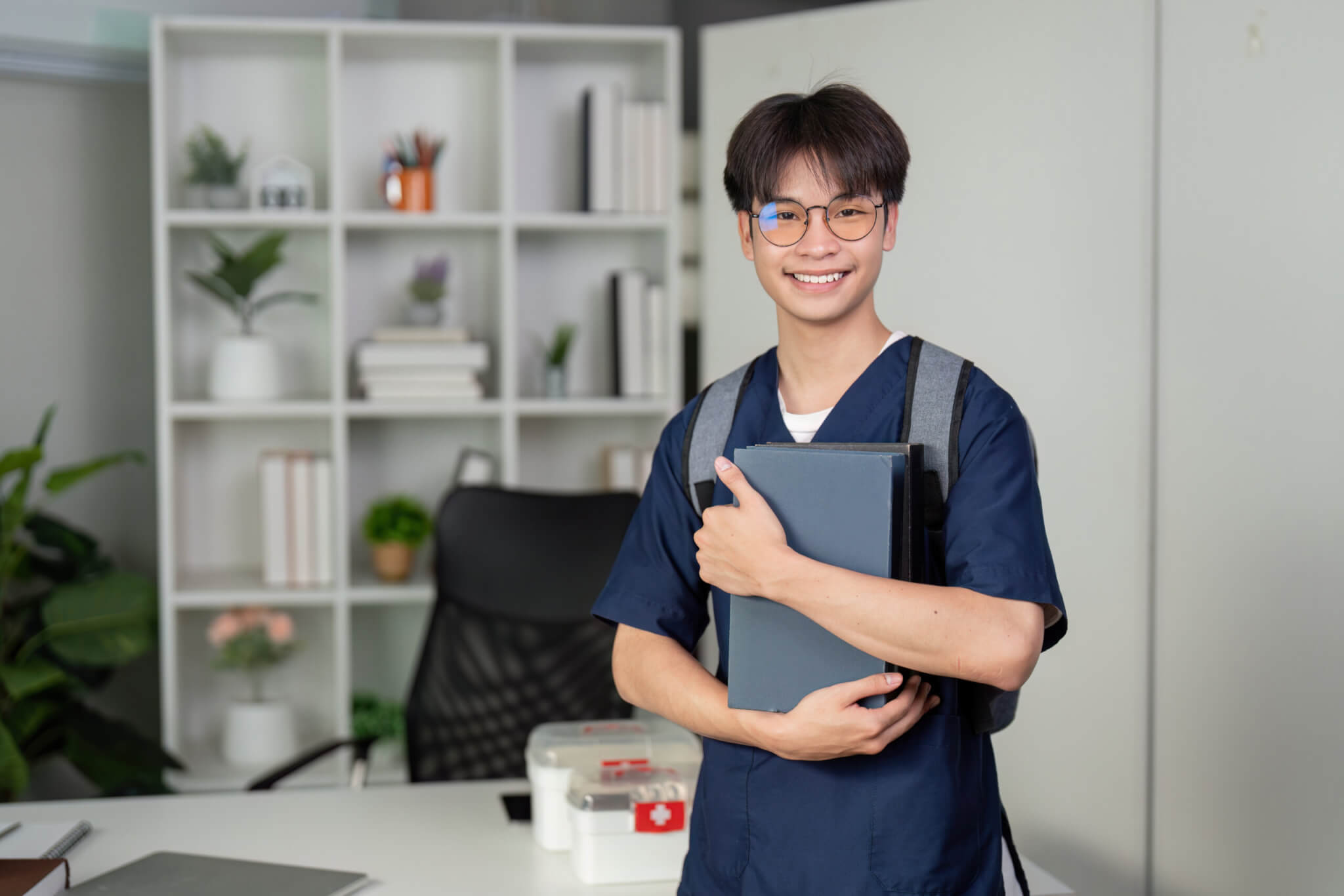 A smiling student in scrubs with a backpack holds books in a modern office with shelves and plants behind him, ready to pursue exciting healthcare careers.