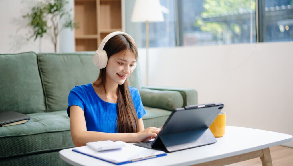 Woman wearing headphones smiling while using a tablet at a coffee table in a bright living room.