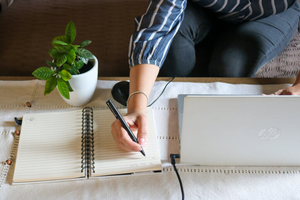 Person writing in a notebook beside a laptop and a potted plant on a table, gathering valuable Labor Market Information for research or analysis.