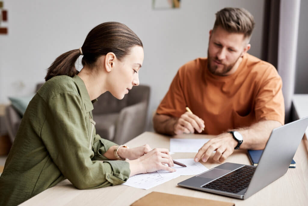 Two people discussing papers while working at a desk with a laptop.
