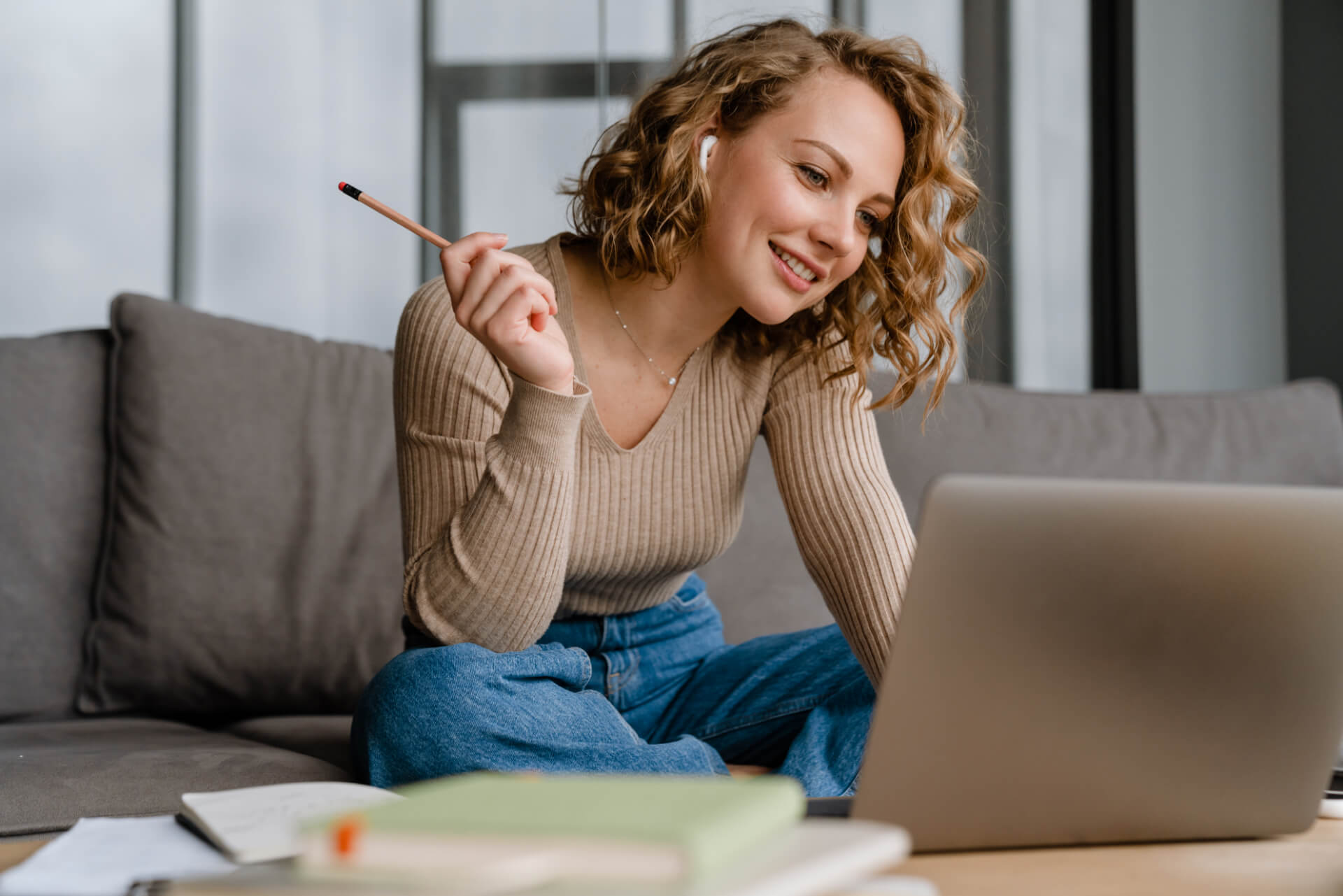 Young woman smiling, sitting on a couch with a laptop, holding a pencil, studying or working from home.